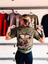 Load image into Gallery viewer, Woman modeling olive green 85th Bike Week 2026 Main Street Station t-shirt showing large vintage back graphic design in Daytona Beach garage setting